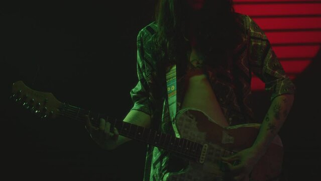 Tattooed rock musician in open shirt playing the guitar in a dark studio with green and red neon illumination. Midsection, zoom shot