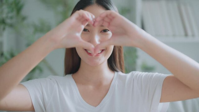 Front view of cute young Asian woman smiling and looking at camera with hands showing heart shape. Facial expressions and positive emotions, healthy life, love and charity