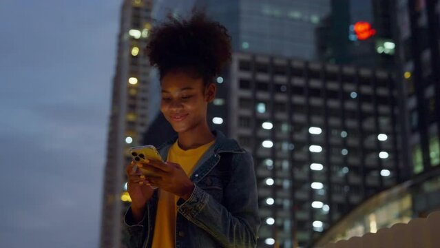 Beautiful young african woman standing in city street at night using mobile smartphone. Happy afro woman typing messages on her mobile phone