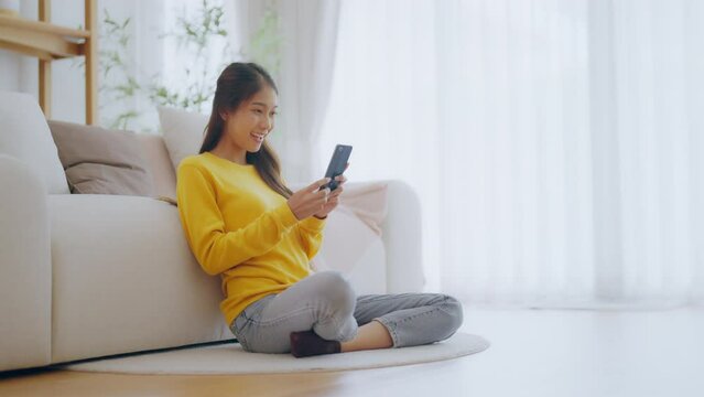 Happy young asian woman relax on comfortable couch at home texting messaging on smartphone, smiling girl use cellphone chatting, online shopping at home, video call communication