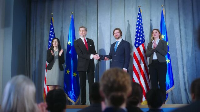World leaders during meeting with journalists. European union representative and mature president of the United States together shake hands after press conference. Backdrop with American and EU flags.