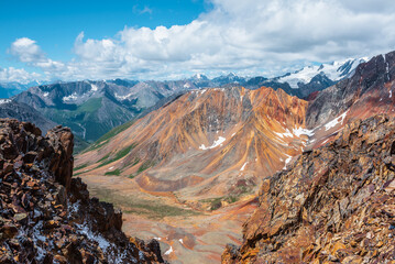 Top view from abyss edge between rocks to multicolor valley with iron river and big sharp rocky ridge of red color. Colorful large mountains in freshly fallen snow in low clouds. Vivid alpine scenery. - 784252108