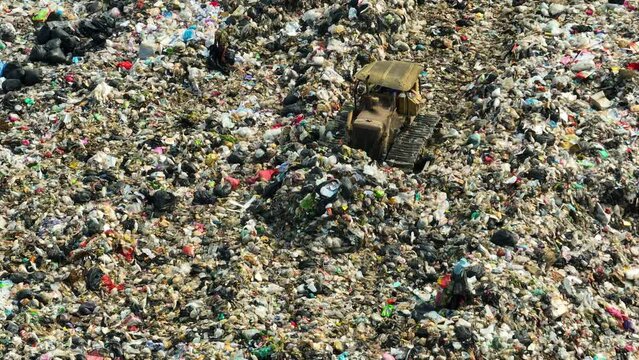From above, a landfill sprawls, a mosaic of waste. A lone bulldozer toils amidst a kaleidoscope of hues, blending plastic, fabric, and assorted refuse in a chaotic dance.
