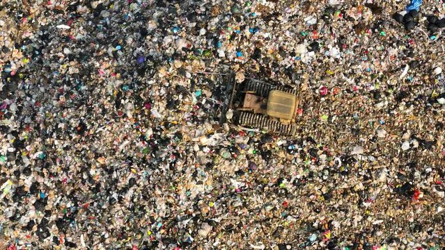 An aerial panorama reveals a landfill's expanse. Amidst a patchwork of colors, a bulldozer maneuvers through a labyrinth of discarded plastics, fabrics, and sundry debris.
