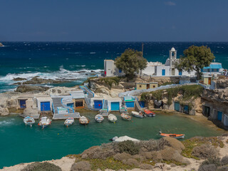 bunte Fischerhäuschen im kleinen Hafen von Mandraki auf der Insel Milos, blau türkises Meer hinter einer kleinen blau weißen Kirche - obrazy, fototapety, plakaty