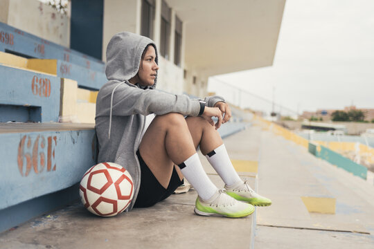 Young woman in gray hoodie sitting with soccer ball on stadium tribune