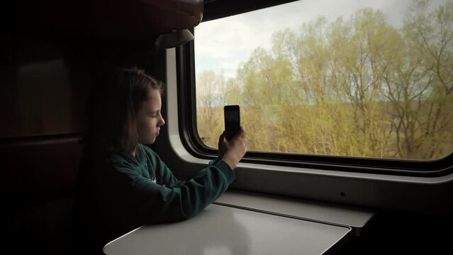 Teenage girl is traveling on a train. A schoolgirl takes pictures of nature on a smartphone through a train window, sitting in the passenger seat at a table. Traveling by train on vacation or moving.