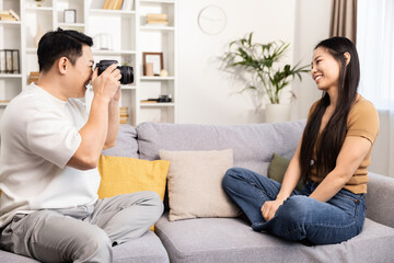 A joyful home setting captures a man taking photographs of a smiling woman sitting comfortably on a couch.