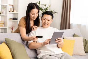 Happy Couple Shopping Online with Credit Card at Home. Cheerful man and woman sitting on sofa, shopping on tablet with credit card in a cozy living room.