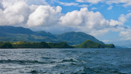 View from the sea to the shore of a tropical island. Mountainous tropical island covered with green jungle, thick cumulus clouds, small waves on the sea surface.