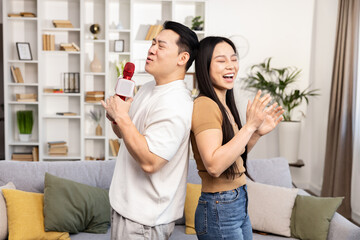 Joyful Asian couple having a fun karaoke session at home. Man sings into a microphone while woman...
