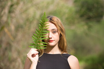 Portrait of a young, beautiful blonde woman dressed in black, half of her face is covered with a green leaf in the forest. The woman is at peace with herself.