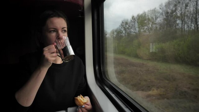 A girl eats a croissant and drinks coffee or tea while sitting at a table in a train compartment and looks out the window at nature. A woman travels by rail.
