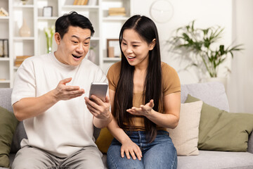 Excited Asian Couple Discovering Fun Content On Smartphone At Home. Candid Shot Capturing Joy, Surprise, And Technology Use In Everyday Life.