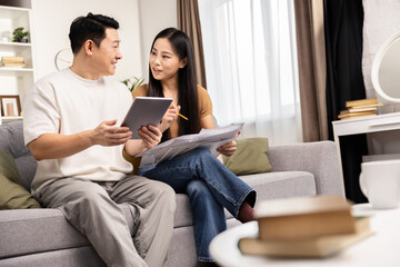 Couple sitting on sofa at home reviewing documents and using tablet, concept of finance, planning, and home life.
