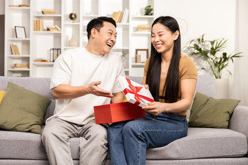 An Asian couple shares a joyful moment exchanging gifts on a cozy sofa, embodying happiness, togetherness, and a warm gift-giving experience.