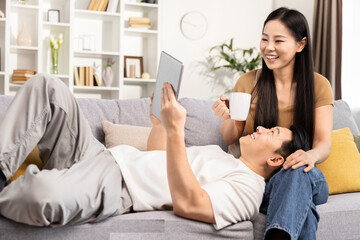 Happy Asian couple relaxing on sofa with tablet and coffee, enjoying weekend technology use at home