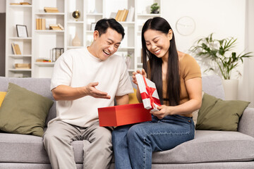 Couple Sharing a Surprise Gift at Home.Happy Asian couple sitting on the couch, enjoying a moment of surprise as the woman opens a gift box with a delighted expression.