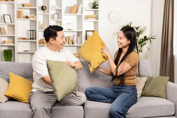 A joyful Asian couple engaging in a playful pillow fight in their cozy living room, laughing and enjoying quality time together.