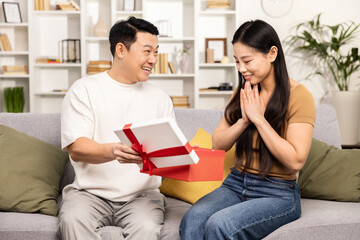 Surprised woman receiving a gift from a man, both sitting on sofa at home, expressing excitement and joy, with a cozy interior background