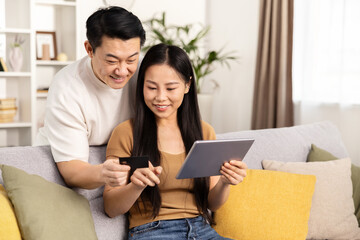 Couple shopping online together at home, holding credit card and tablet, smiling, engaging in e-commerce