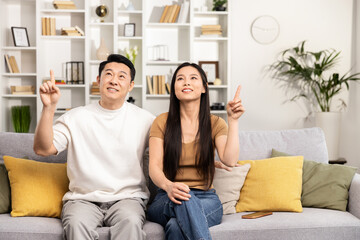 Happy Asian couple sitting on a couch in a cozy living room, pointing upwards with smiles, enjoying time at home.