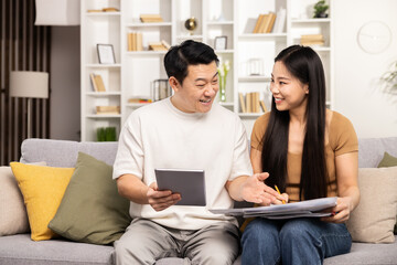 Asian Couple Planning On Couch With Tablet And Documents. Joyful, Engaged Asian Man And Woman Discussing Finances Or Plans At Home, Creating A Relaxed Yet Focused Environment.