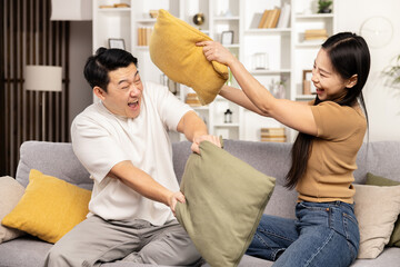 A joyful couple engages in a playful pillow fight on their cozy couch in a well-lit living room, sharing a fun, carefree moment together.