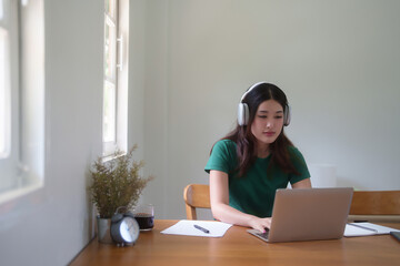 Young asian student women wear headphone to studying and watching lecture webinar in online class on laptop while typing to researching information and learning knowledge remote education from home - 784334952