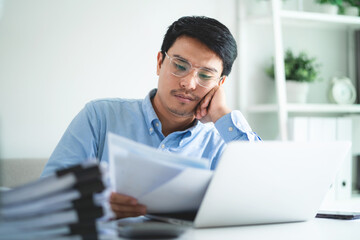 A man is sitting at a desk with a pile of papers in front of him. He is looking at a laptop and he is tired or stressed