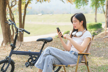 Young asian women checking social media entertainment and reading message chatting on smartphone while sitting on chair to relaxation with journey travel lifestyle after cycle bicycle in the park - 784336584