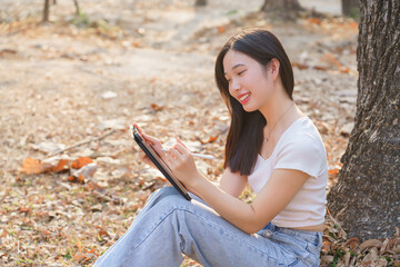 Young asian women sitting on the grass under tree in the nature park and using digital tablet to writing business information while working outside and relaxation with journey travel lifestyle - 784337328