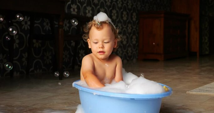 In basin filled with warm water and white foam, pensive little boy sits calmly. With downcast gaze, he remains oblivious to soap bubbles flying in front of him. Scene unfolds in slow motion