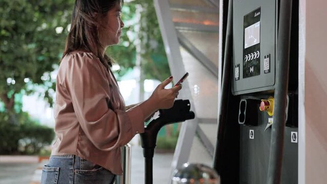 Business asian woman using smartphone electric car recharge battery at downtown charging point clean emission transport