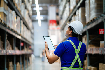 A man in a blue shirt and a white hat is looking at a tablet while standing in a warehouse