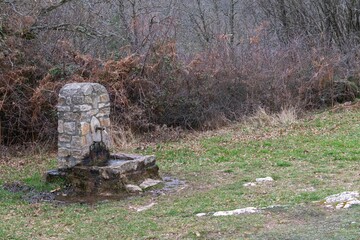 Small ancient fountain made from stones on grassland and autumn plants behind it