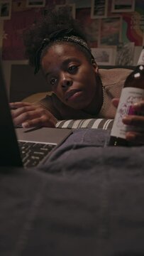 Vertical shot of young tired African American woman with glass bottle of beer in hand awaking from sleep on bed beside laptop while spending weekend night alone in her bedroom