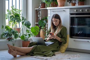 Thoughtful woman with laptop on knees looking at screen cellphone sits comfortably on floor among...