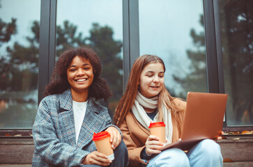 Two happy multiracial teen girls looking at laptop and laughing while drinking coffee in autumn...