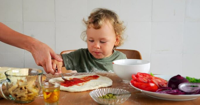 Young boy is learning to make homemade pizza, scooping tomato sauce with spoon, his mom takes his hand and shows him how to spread sauce on dough. Little one is more interested in tasting everything