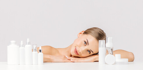 Obraz na płótnie Canvas Nice woman on a white background among jars, bottles and tubes for cosmetic procedures. Production of natural cosmetics. Cosmetics based on natural ingredients, scrub, tonic, body care.