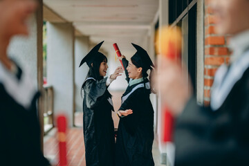 At the graduation ceremony, two Asian university students in gowns happily embrace, wishing each...