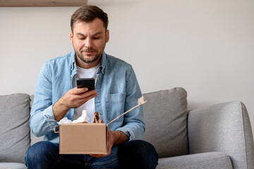 Man in a denim shirt unpacks a package and takes a photo of his purchase, internet order. Person sharing feedback with online store using his smartphone. - obrazy, fototapety, plakaty