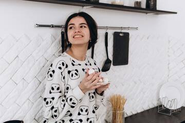 Cheerful woman standing in bright kitchen smiling eat candy, dressed in patterned pajama set. The...