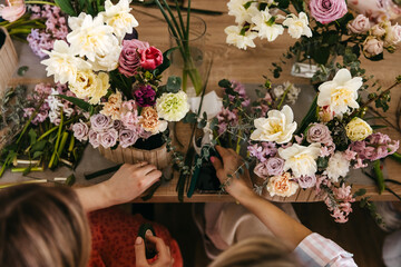 Overhead view of hands crafting floral arrangements with a rich mix of daffodils, roses, and...