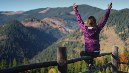 Tourist woman enjoys her morning in the mountains. Girl sitting on fence raises her hands up...