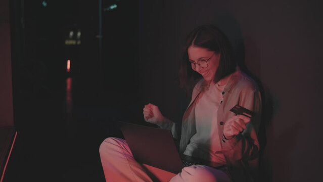 Woman sits on the floor in a dark room with red dim lights and uses a laptop. The girl enters her card details to pay for the goods and is happy. A beautiful young woman in glasses communicates