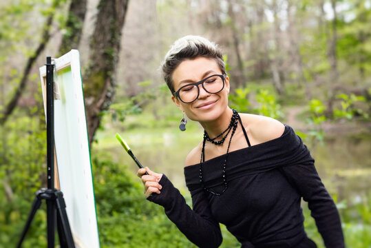 A beautiful girl artist with glasses poses sensually and emotionally against the background of a forest with brushes and paints in her hands experiencing happiness