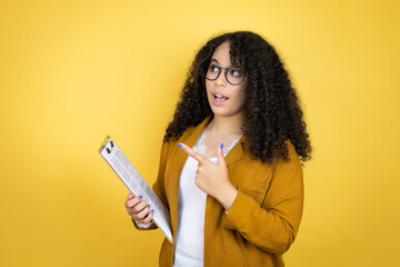 African american business woman with paperwork in hands over yellow background amazed and pointing...