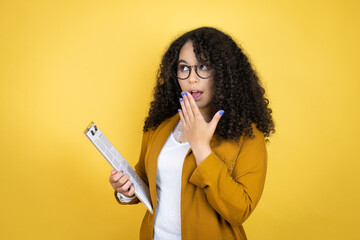 African american business woman with paperwork in hands over yellow background covering mouth with hand, shocked and afraid for mistake. surprised expression